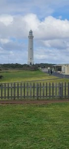 A tall white lighthouse, known as Cape Leeuwin Lighthouse, stands on a grassy hill under a cloudy sky in Augusta. People walk on a paved path nearby, with a picket fence in the foreground.
