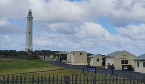 A coastal scene near Augusta features the majestic Cape Leeuwin Lighthouse on the left with a cluster of small buildings on the right. Two people walk along a path under a partly cloudy sky. A wooden fence runs in the foreground, marking the start of a scenic Trike Tour route.