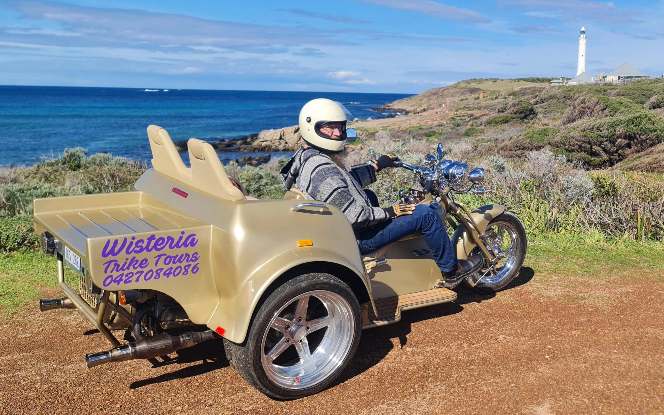 A person wearing a helmet and jacket sits on a gold trike overlooking a coastal view with a lighthouse in the background. The trike features 