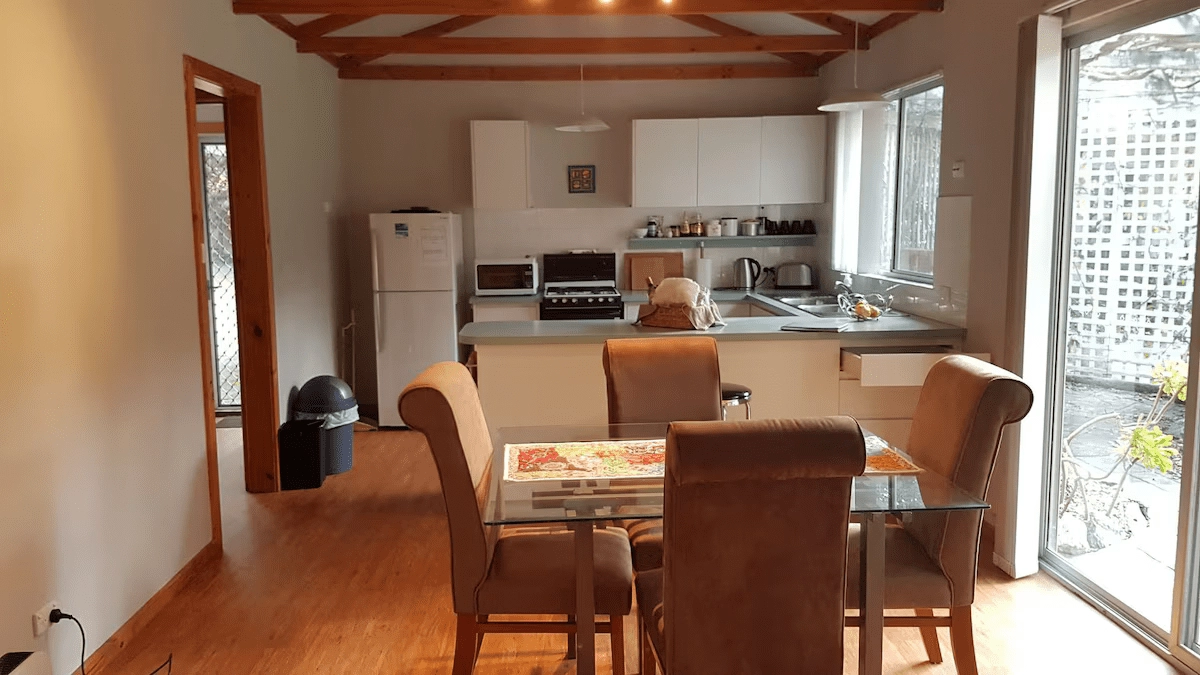 A modern kitchen and dining area with wooden floors, brown cushioned chairs, a glass dining table, white cabinetry, a refrigerator, and a stove. Large windows provide natural light.