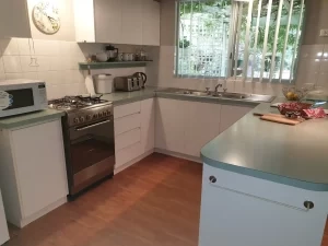 A clean, modern kitchen with a gas stove, white cabinets, microwave, kettle, toaster, and a large window above the sink. A cutting board with snacks sits on the countertop.