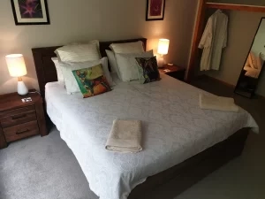 A neatly made bed with patterned pillows and folded towels at the foot. Two bedside tables with lamps sit on either side, and an open closet reveals a hanging white robe.