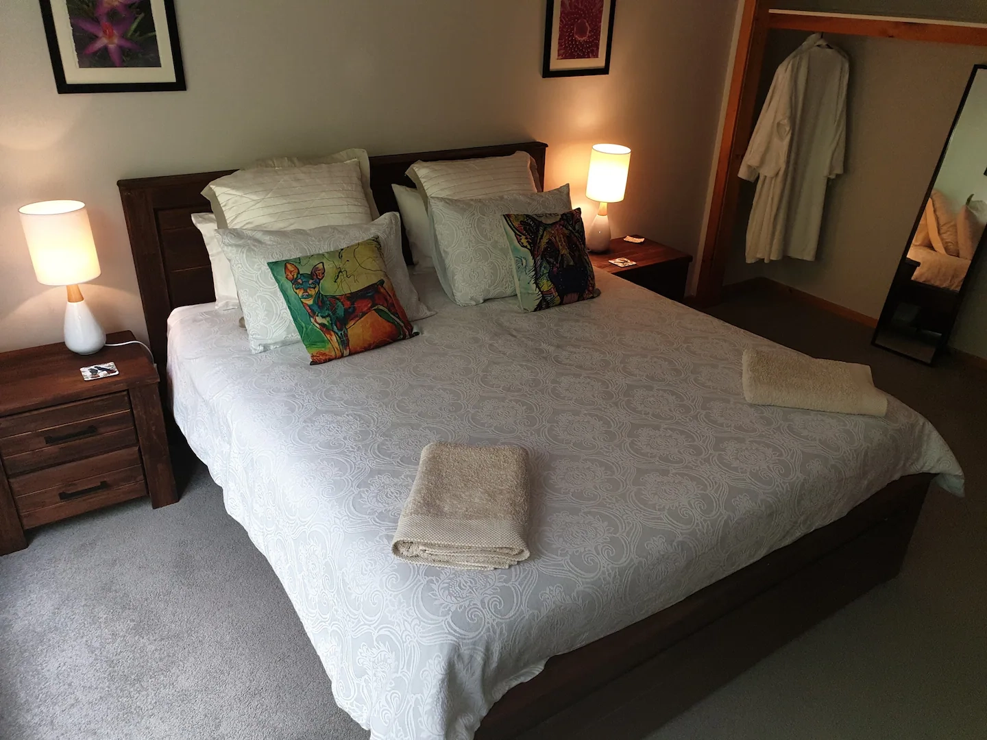 A neatly made bed with patterned pillows and folded towels at the foot. Two bedside tables with lamps sit on either side, and an open closet reveals a hanging white robe.
