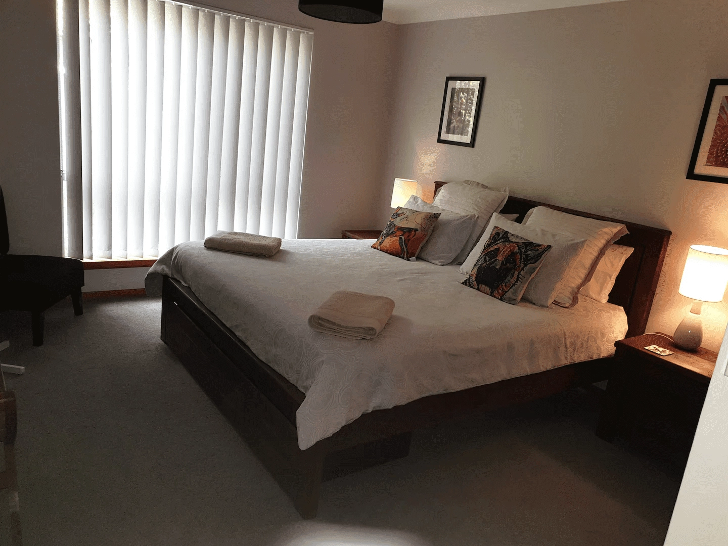 A bedroom with a large bed covered in white linens, two rolled towels, two bedside tables with lamps, framed pictures on the walls, and vertical blinds covering a window to the left.