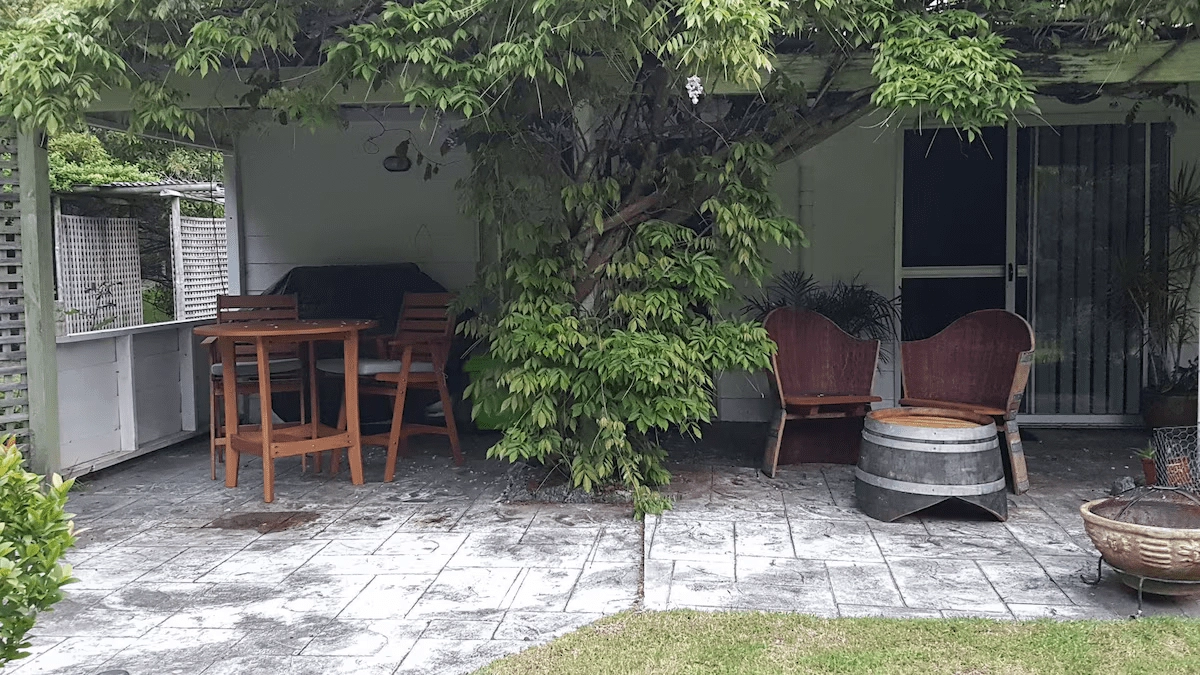 A patio area with a wooden table and chairs under a tree. There are two wooden tub chairs and a barrel table nearby. The area has a tile floor and a sliding glass door in the background.