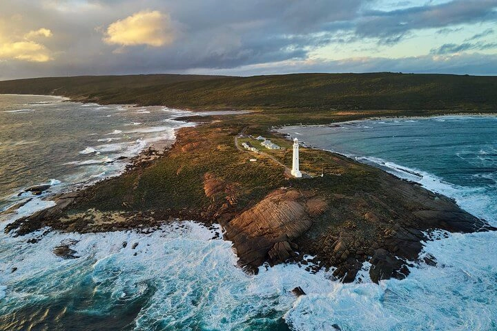 Aerial view of a lighthouse on a rocky peninsula surrounded by ocean waves, adjacent to a quaint cottage with a grassy landscape and cloudy sky in the background.