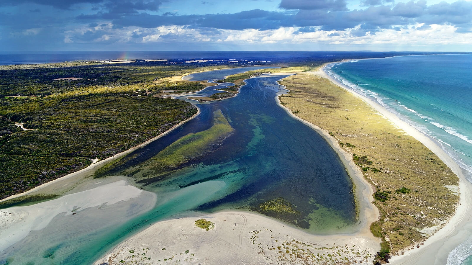 Aerial view of a coastal landscape with a river flowing into the ocean, surrounded by sandy shores, grassy areas, and dense vegetation under a partly cloudy sky, with a charming cottage nestled among the greenery like something out of a book.