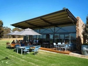 Hamelin Bay Winery Outdoor seating area with tables, chairs, and umbrellas on a patio next to a modern building with large glass windows. Several people are seated and socializing while someone reads a book. A large grassy area in the foreground adds a touch of cottage charm.