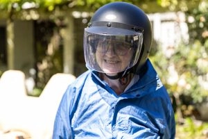 A Person Wearing Glasses And A Blue Jacket, Smiling While Wearing A Black Helmet With A Clear Visor, Standing Outdoors.