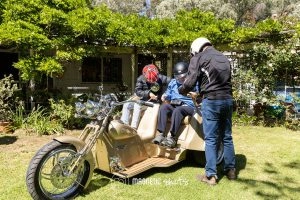 Three People Prepare To Ride On A Three Wheeled Motorcycle In A Garden, With Two Seated And One Standing. All Wear Helmets, Surrounded By Green Foliage And A Small Building In The Background.