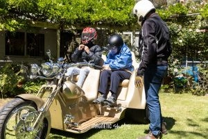 Two People Sitting In A Sidecar Attached To A Motorcycle, Adjusting Their Helmets. A Standing Person In A Helmet Talks To Them. Surrounded By Greenery.