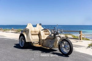 A Beige Three Wheeled Motorcycle Parked On A Coastal Road With The Ocean In The Background Under A Clear Blue Sky.