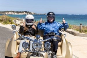 Two People Wearing Helmets Sit In A Motorcycle Sidecar On A Coastal Road, Giving Thumbs Up With The Ocean And Rocky Landscape In The Background.