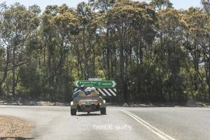 A Small Vehicle Is On A Rural Road Approaching A Signpost Indicating Directions To Margaret River And Augusta, With Trees In The Background.
