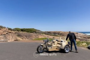A Person Stands Beside A Beige Three Wheeled Motorcycle On A Coastal Road With Rocky Terrain Under A Clear Blue Sky.