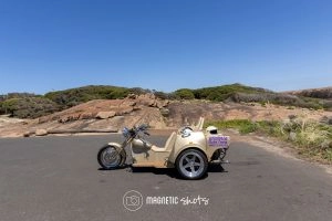 Three Wheeled Yellow Motorcycle Parked On A Coastal Road With Rocky Terrain And Clear Blue Sky In The Background.