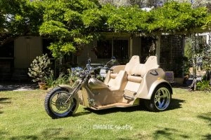 A Beige Three Wheeled Motorcycle Parked On Grass In Front Of A Lush Garden Setting, With A Two Seat Passenger Area.