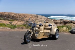 A Beige Three Wheeled Motorcycle Parked On A Coastal Road With Rocky Shoreline And Ocean Waves In The Background.