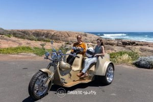 Two People Are Seated On A Trike Parked On A Coastal Road With Rocky Terrain And The Ocean In The Background.
