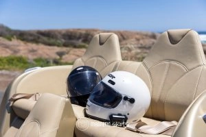 Two Motorcycle Helmets, One White And One Black, Rest On The Beige Seats Of An Open Top Car With A Rocky Landscape In The Background.