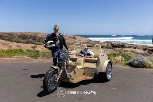 A Person Stands Next To A Cream Colored Three Wheeled Motorcycle On A Coastal Road With Rocky Terrain In The Background.