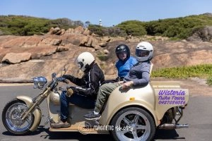 Three People Wearing Helmets Ride A Cream Colored Trike On A Scenic Road With Rocks And Greenery In The Background.
