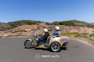 Two People Wearing Helmets Ride A Beige Trike With A Sign For Wisteria Tours, Parked On An Asphalt Surface With Rocky Hills And Clear Blue Sky In The Background.