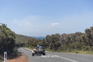 A Three Wheel Vehicle Drives On A Scenic Road Surrounded By Trees, With The Ocean Visible In The Background Under A Clear Sky.