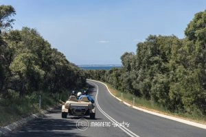 A Small Dune Buggy Travels Down A Winding, Tree Lined Road Toward The Coast On A Clear Day.