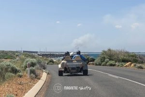 Two People Drive A Dune Buggy On A Coastal Road, With Ocean And Smoke In The Distant Background Under A Clear Sky.