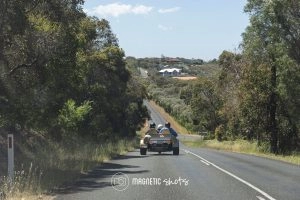 A Vintage Car Drives Down A Rural Road Lined With Trees, With Hills And Houses Visible In The Background.