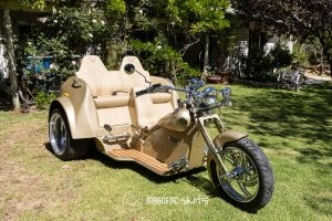 A Custom Beige Three Wheeled Motorcycle With A Two Seater Sidecar Is Parked On A Grassy Lawn, With Trees And A House In The Background.