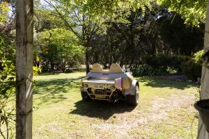 A Small Custom Car With Two Seats And An Australian License Plate Is Parked On A Grassy Area Surrounded By Trees.